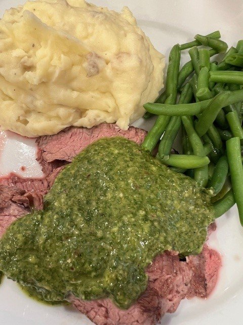 Picture of flank steak on a plate with mashed potatoes, green beans, and chimichurri sauce.