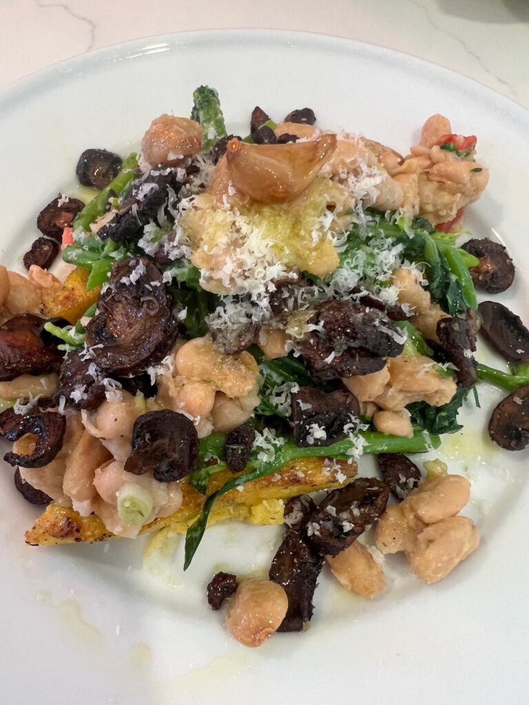 Picture of grilled polenta on a white plate topped with white beans, broccoli rabe, diced tomatoes, garlic, and grilled baby bella mushrooms.