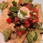 Picture of a plate of nachos topped with ground meat, black beans, corn, tomatoes, guacamole, sour cream and green onions.