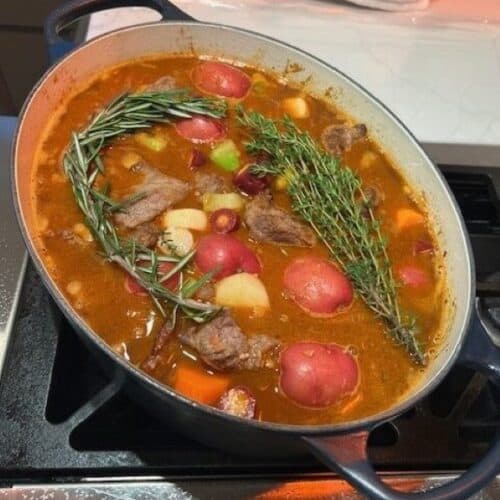 Picture of a Classic Beef Stew cooking on the stove in a navy blue Dutch oven topped with rosemary and herbs.