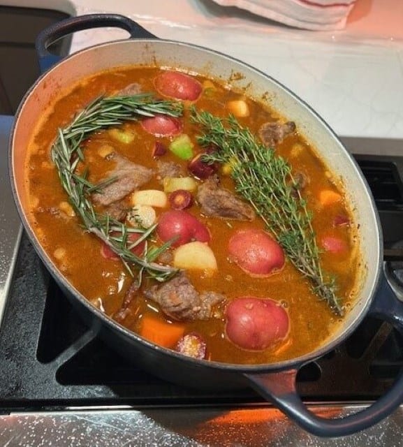 Picture of a Classic Beef Stew cooking on the stove in a navy blue Dutch oven topped with rosemary and herbs.