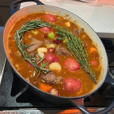 Picture of a Classic Beef Stew cooking on the stove in a navy blue Dutch oven topped with rosemary and herbs.