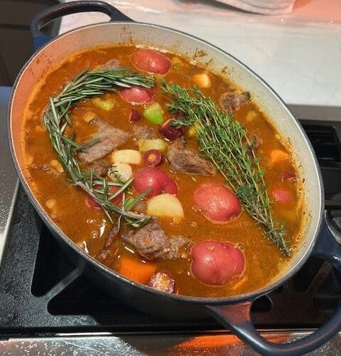 Picture of a Classic Beef Stew cooking on the stove in a navy blue Dutch oven topped with rosemary and herbs.