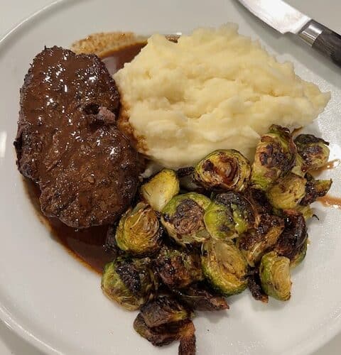 Picture of a filet mignon on a white plate with garlic mashed potatoes and roasted Brussels sprouts.