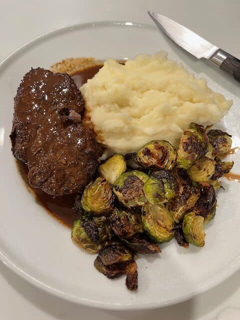 Picture of a filet mignon on a white plate with garlic mashed potatoes and roasted Brussels sprouts.