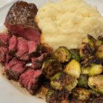 Picture of sliced filet mignon on a white plate with garlic mashed potatoes and roasted Brussels sprouts.