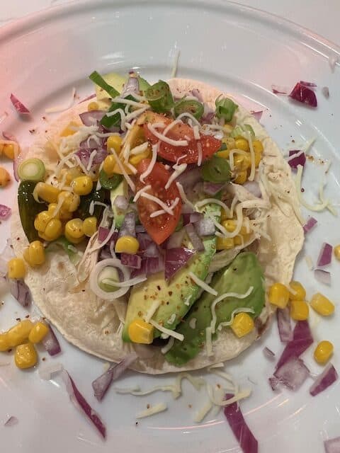 Picture of an open chicken taco topped with black beans, avocado, corn, onions, tomato, and cheese.