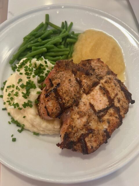 Picture of a juicy bone-in pork chop on a white plate paired with mashed potatoes, green Beans and applesauce.
