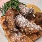 Picture of golden waffles on a white plate topped with crispy chicken wing and paired with green beans and applesauce. The dish is topped with maple syrup and powdered sugar.