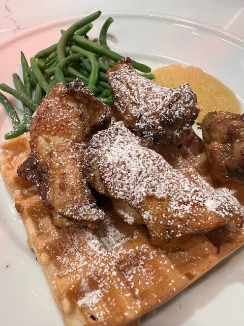 Picture of golden waffles on a white plate topped with crispy chicken wing and paired with green beans and applesauce. The dish is topped with maple syrup and powdered sugar.