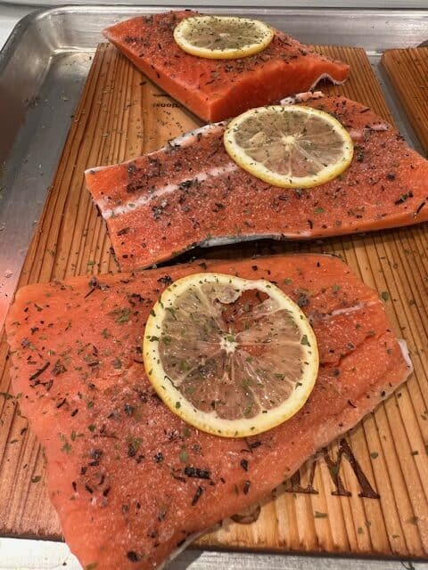 Picture of raw salmon filets on a cedar plank board with a slice of lemon on top.